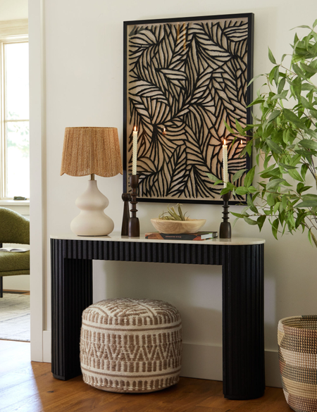 Entryway vignette featuring a fluted wood entryway table, patterned ottoman, table lamp with a woven shade, and botanically-inspired artwork