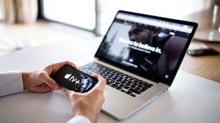 Hands hold smartphone with Apple TV Plus logo, next to a MacBook with Apple TV Plus also on it