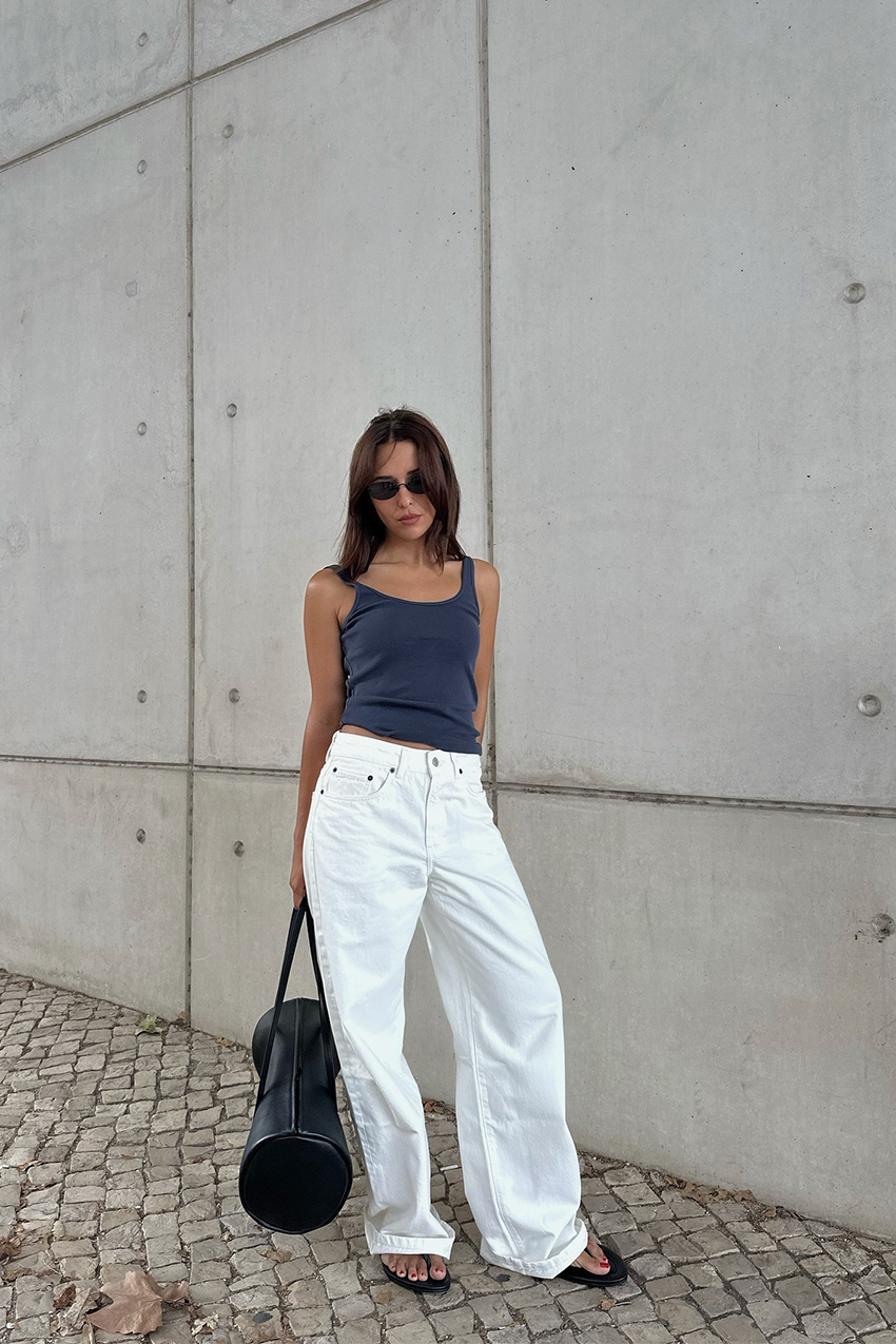A woman standing outside in front of a concrete wall wearing a navy blue tank top with baggy white wide-leg jeans, black flip-flops, black sunglasses, and a black large barrel bag.