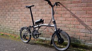 The bottle-shaped Cytronex e-bike conversion kit fitted to the top tube of a Brompton, leaning against a brick wall
