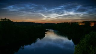 Silvery noctilucent clouds are pictured shining above the Neris River in Vilnius, Lithuania as an orange sunset fades near the horizon.