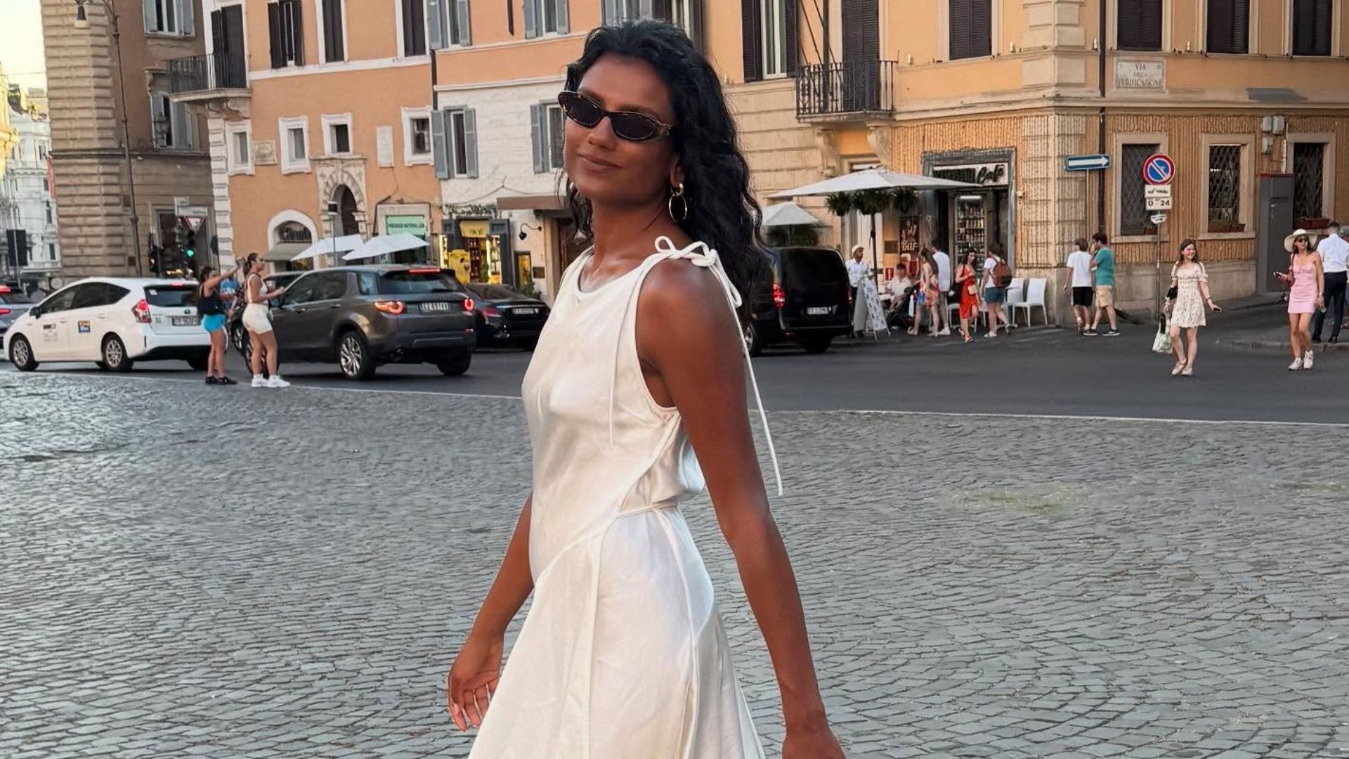 Simone Ashley wears a white lace-trim dress with black sandals and a tan bag as she walks through the streets in Italy.