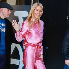 Gwyneth Paltrow steps outside wearing a pink satin trouser and shirt set with pink high vamp heels.