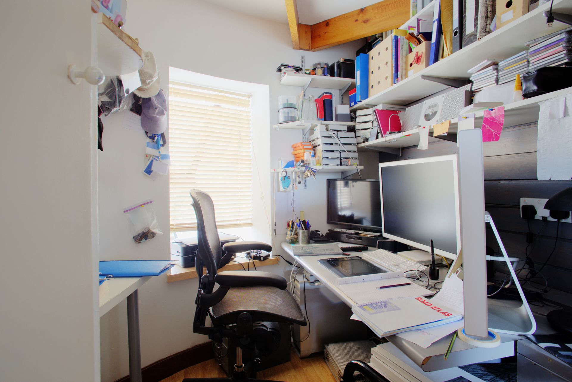 A shot of a messy desk in a home office, the room is small and cluttered, on the desk is three computer monitors and office supplies