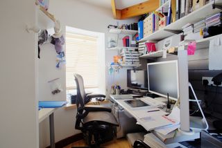 A shot of a messy desk in a home office, the room is small and cluttered, on the desk is three computer monitors and office supplies