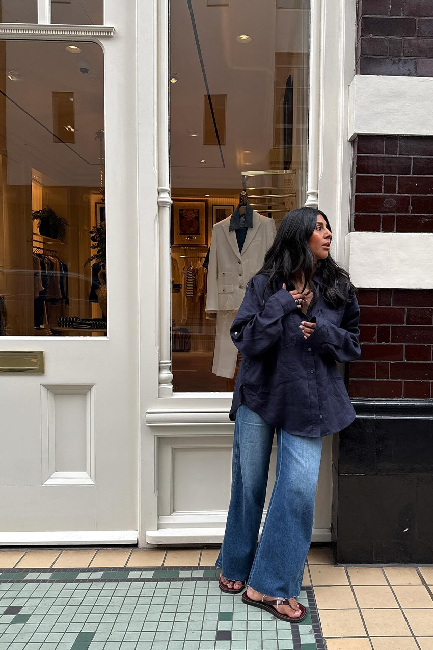 Denim and shoe combos shown in a street style photo of a woman leaning against a storefront outside, wearing an oversized navy button-down shirt with dark wash wide-leg jeans with dark brown flip-flops