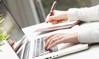 Close up of left hand on a laptop computer keyboard while right hand holds pen