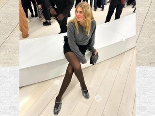 Danish fashion influencer Sidsel Alling sitting at the Loewe fashion show in Paris wearing a gray polo sweater, black feather mini skirt, brown sheer tights, and studded Alaia Mary Jane flats