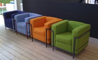 Light wood floor, row of four fabric armchairs with dark grey metal frame and legs, in colours, dark blue, light blue, orange and green, black divider screen, blurred view of room in the distance