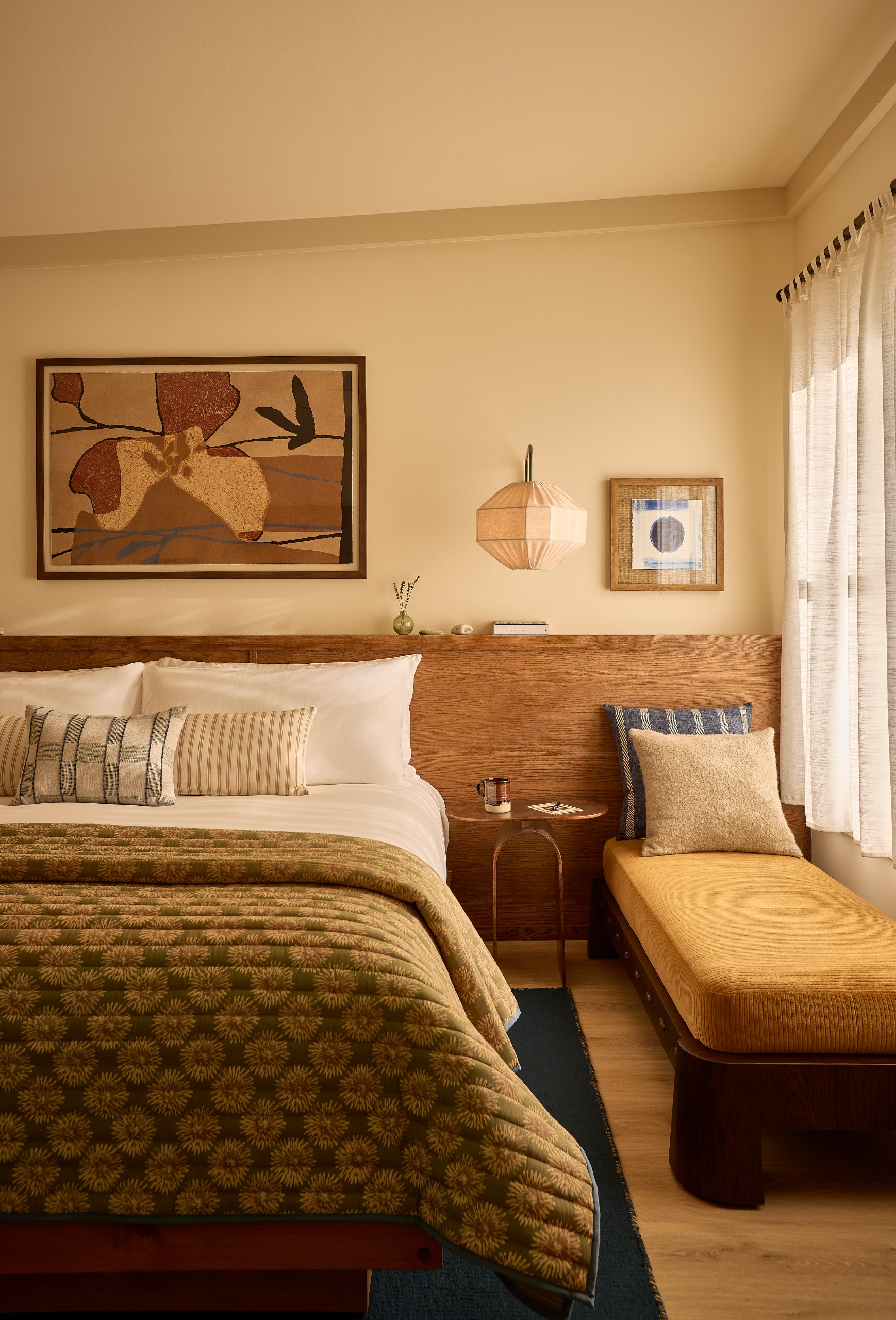 Interiors of a warmly furnished bedroom with ochre, brown, and terracotta tones, a king bed topped with an olive green runner, abstract artwork, warm lighting, and a window seat banquette.