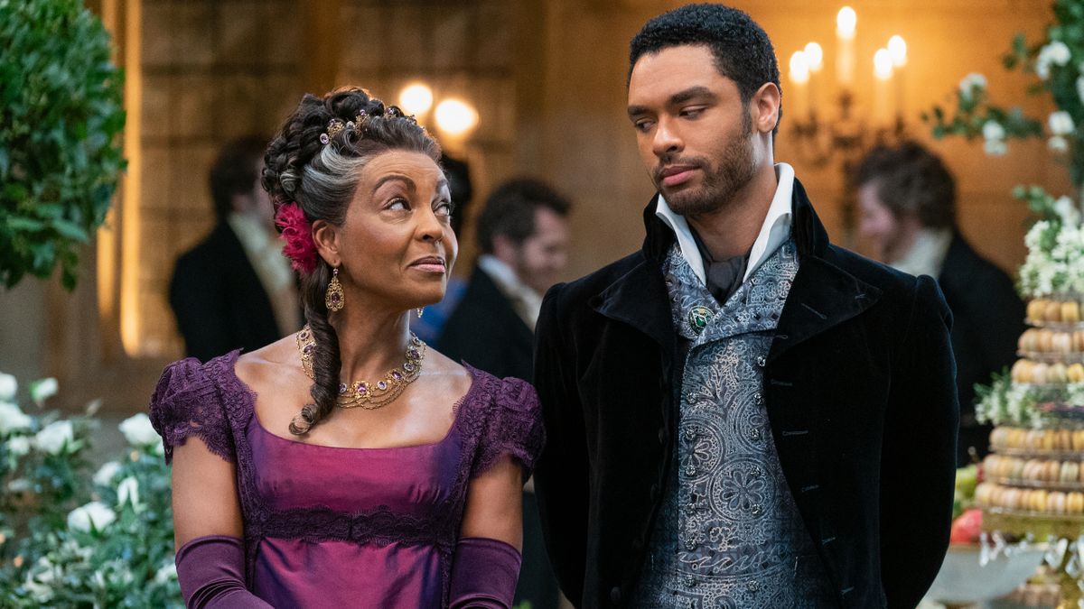 Adjoa Andoh and Regé-Jean Page look slyly at one another at a party in Bridgerton.