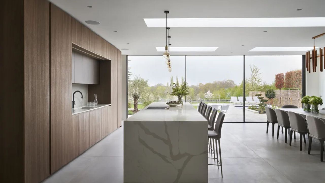 A kitchen with an island in the middle and bar stools beside it. There is also a dining table in the kitchen with plants on top. 