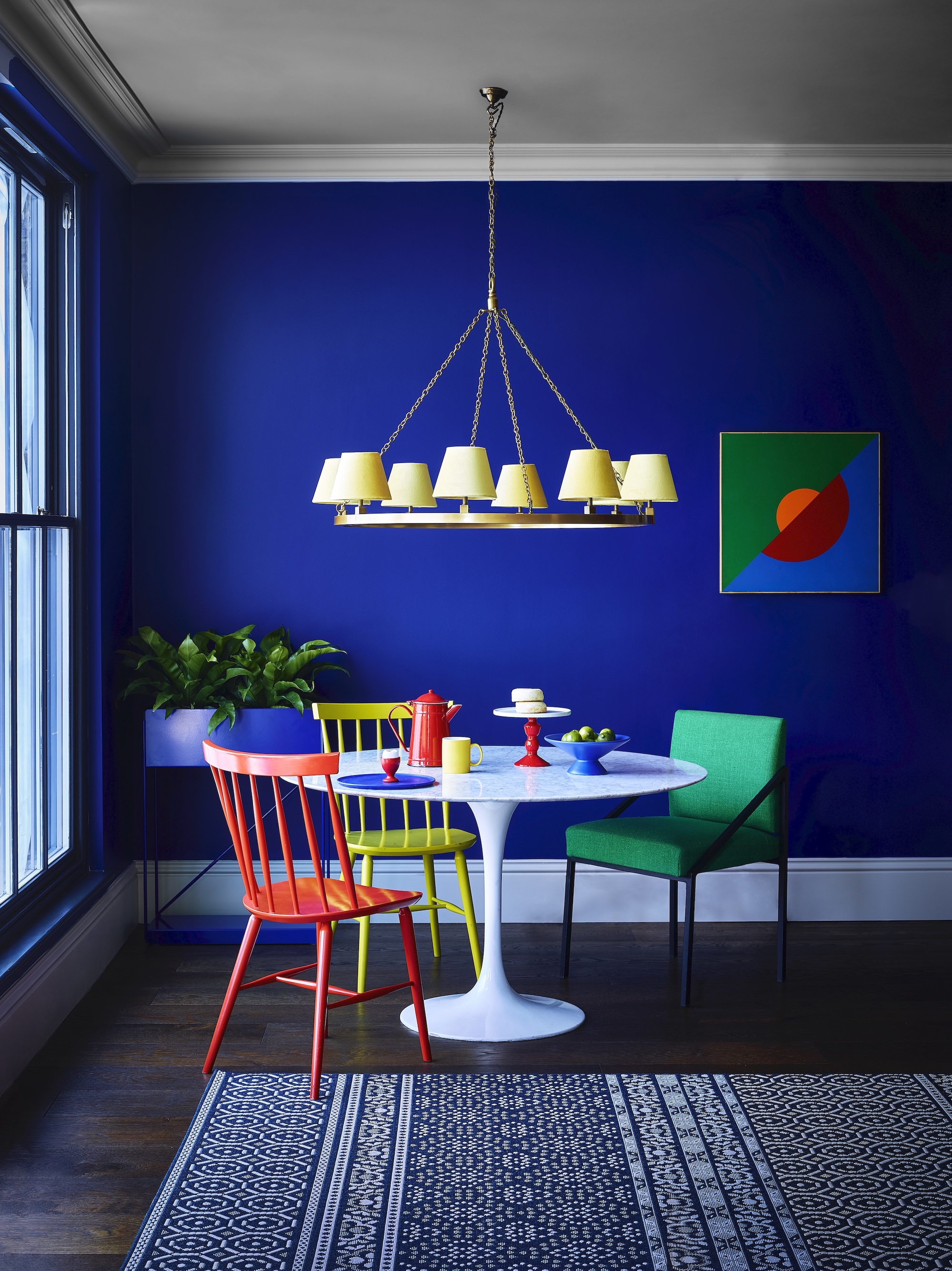 A cobalt blue dining space with dark wood floors and a brightly coloured dining table with miss-matched chairs, above hangs a yellow chandelier.