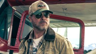 Brad Pitt looking rugged in a tan jacket, green and white ball cab and sunglasses, while standing next to a plane in Heart of the Beast. 