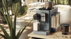 De'Longhi Rivelia fully automatic bean-to-cup coffee maker in pebble grey on light wood countertop with additional hopper