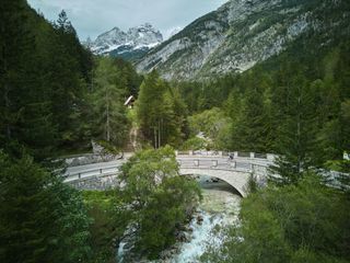 A road cyclist crosses over a river on a stone bridge in Slovenia. Snow capped mountains peak out from behind the trees in the background