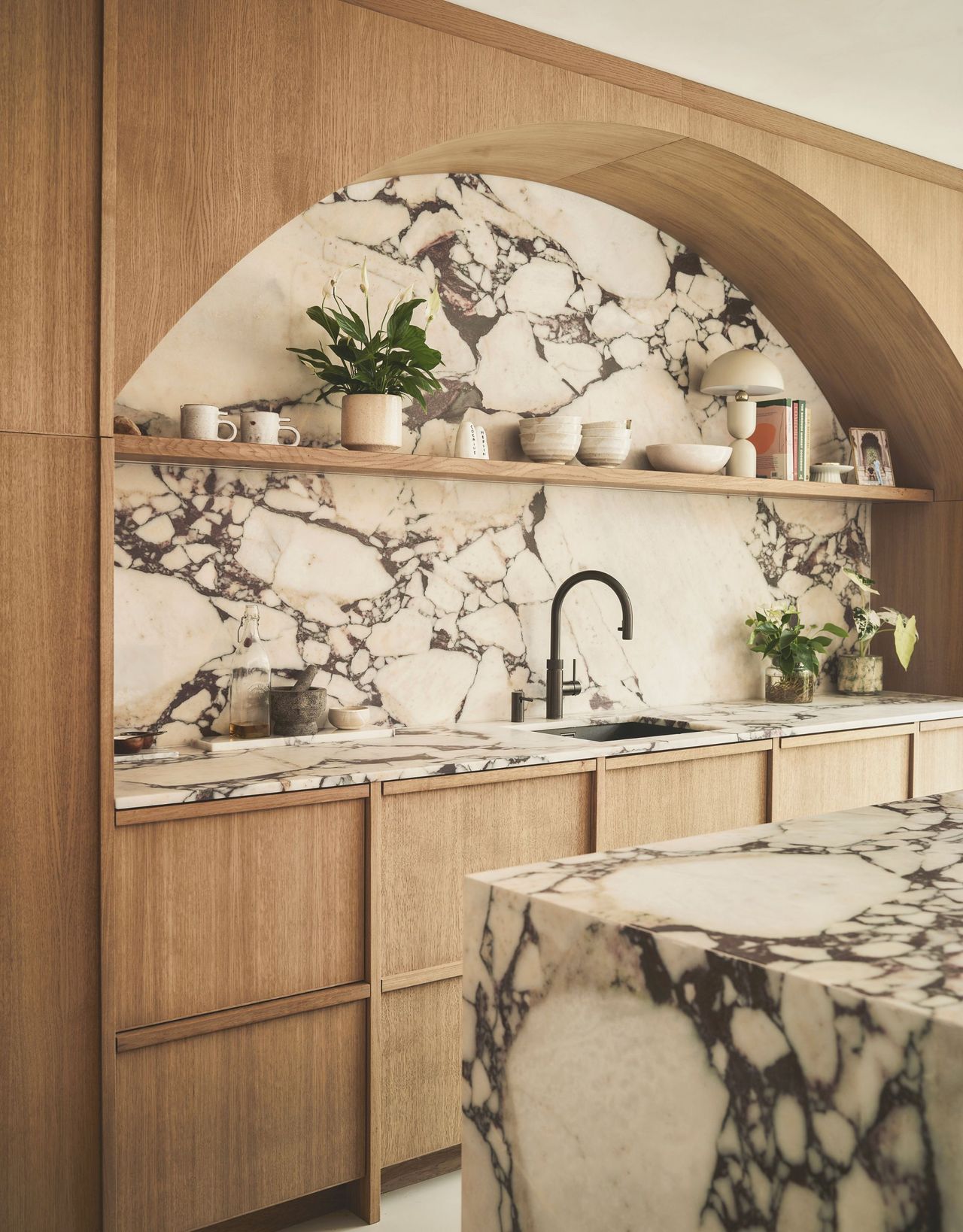 A modern marble and wood kitchen with an arched wood design above a marble backsplash with open shelving and a marble island