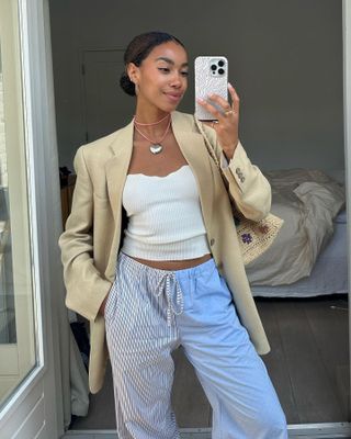 Dutch female fashion influencer Amaka Hamelijnck poses for a mirror selfie wearing a pink cord necklace with a heart pendant, neutral linen blazer, sweetheart neckline top, and blue striped pajama pants.