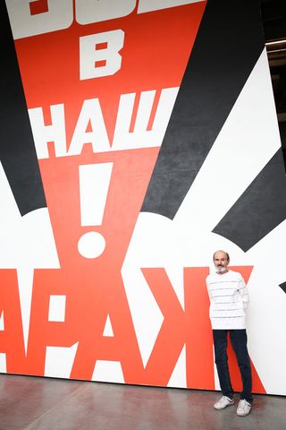 Artist Eric Bulatov standing next to large painted wall which reads 'Come to Garage'