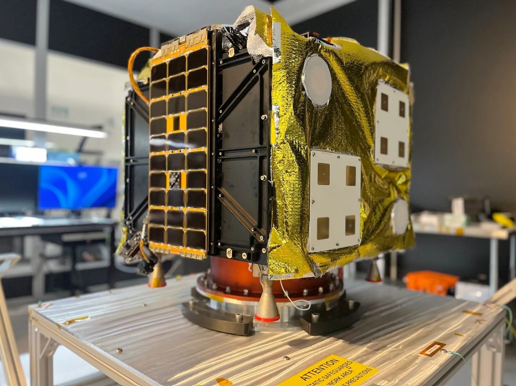 A cube-shaped spacecraft sits secured to a metal table.