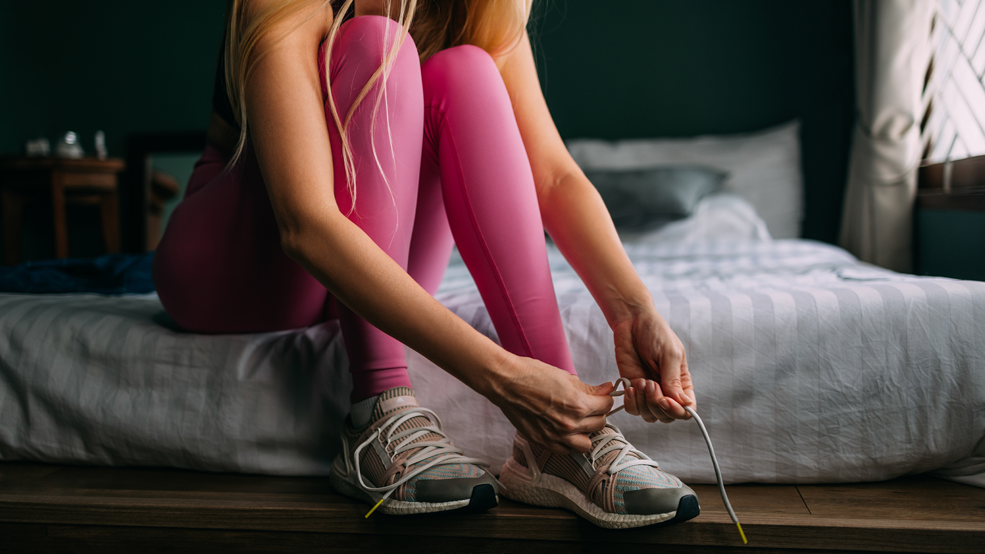 A woman wearing pink leggings sits on the edge of her bed, bending down to tie up her sneaker shoelaces before going out on a run