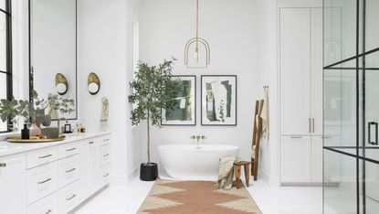 White bathroom with white countertop, white freestanding bath, potted tree, terracotta rug and brass wall sconce and ceiling pendant