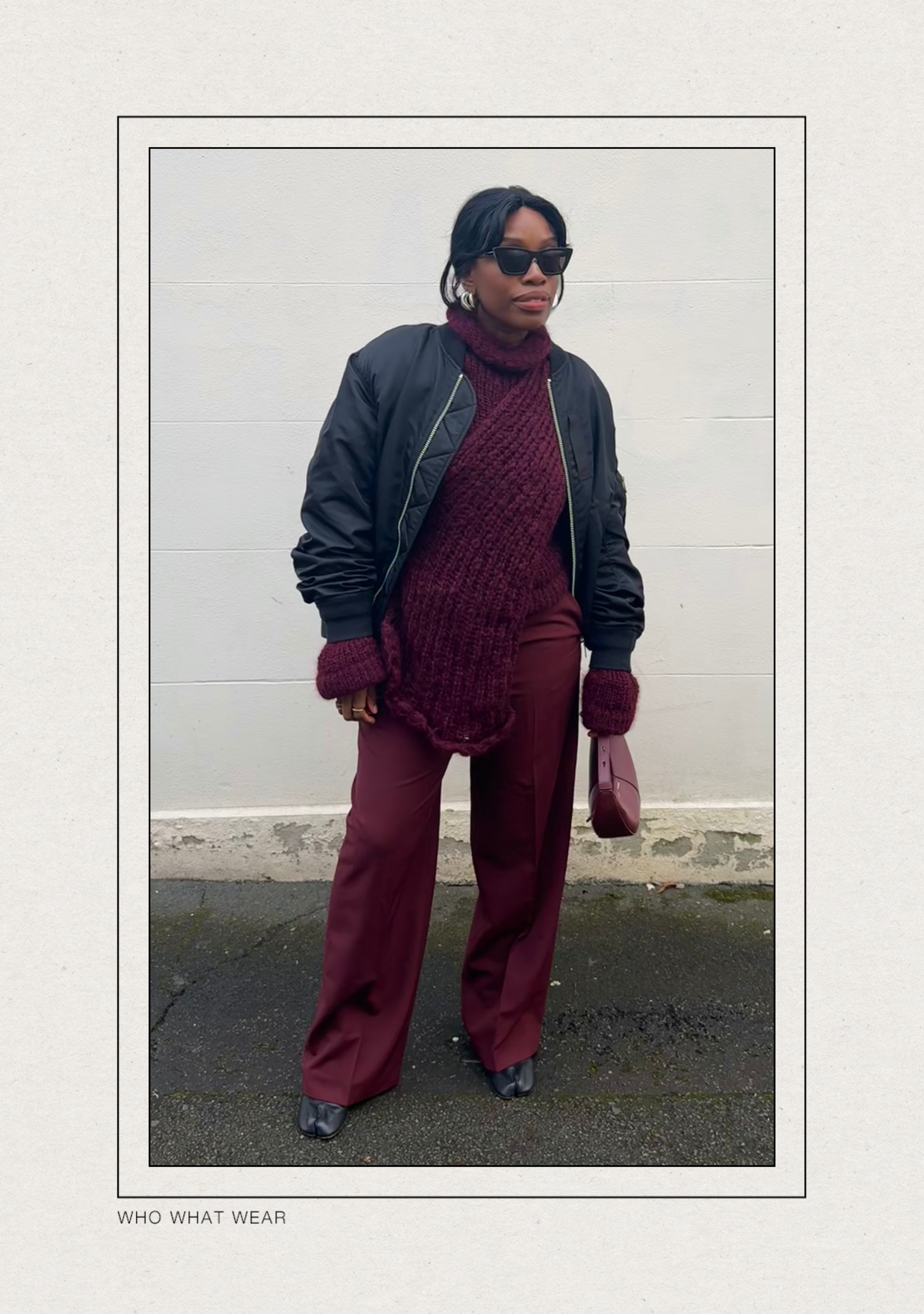 @anuogugbesan wears a black bomber jacket with a burgundy chunky knit jumper, burgundy tailored trousers, Margiela Tabi boots and a burgundy Coperni shoulder bag whilst standing in the street