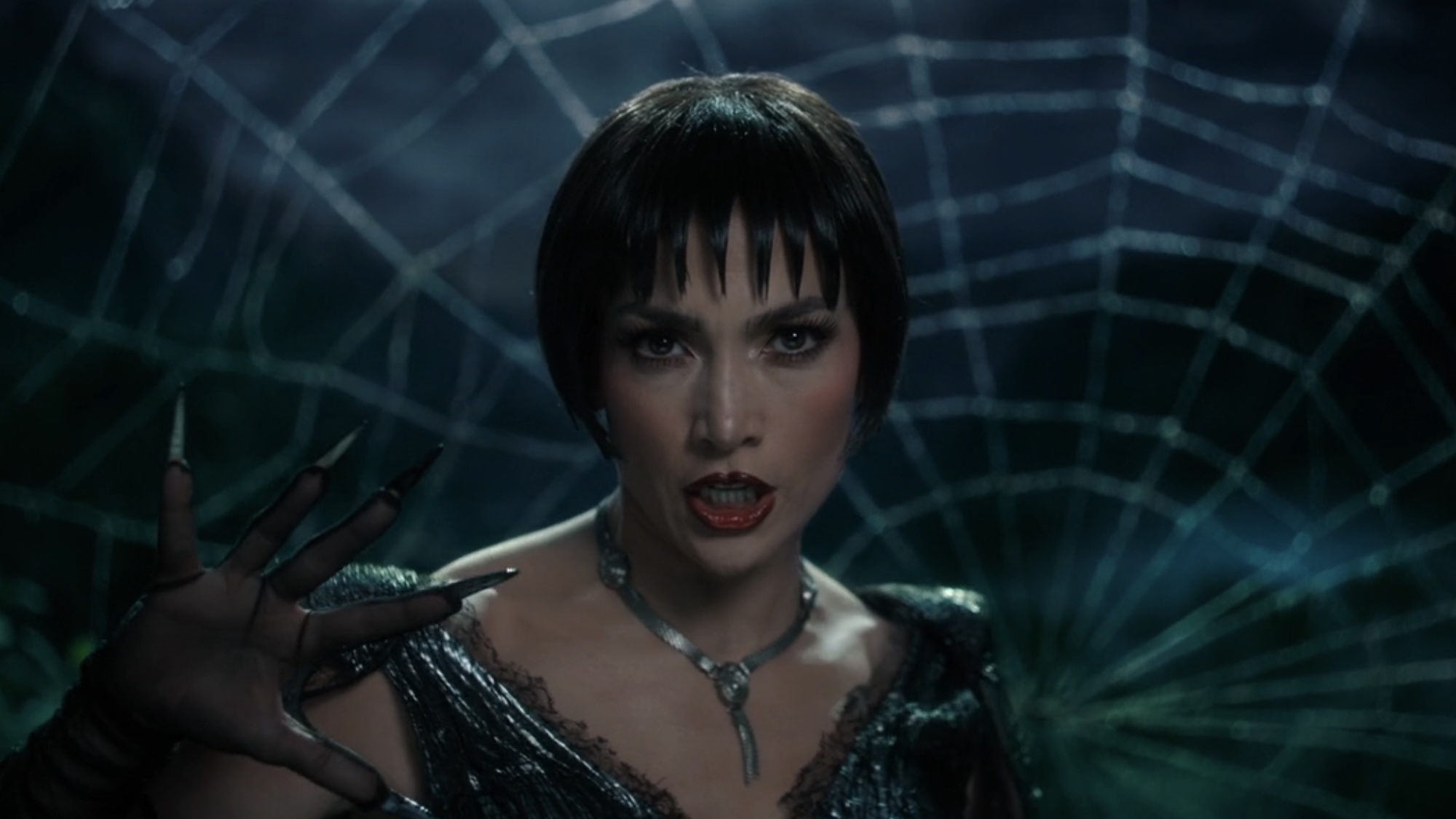 JLo wearing all black in front of a web singing the title song of 'Kiss of the Spider Woman'