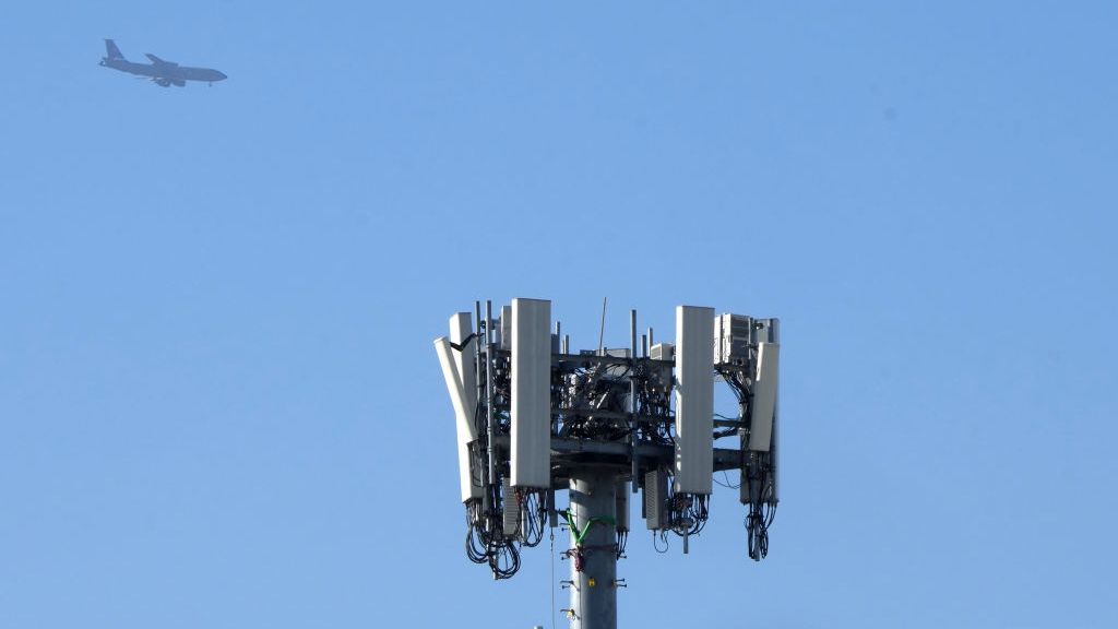 A 5G mast in the foreground, with a clear blue sky and a commercial airliner flying in the distant background