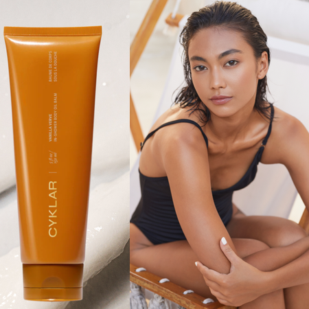 A woman wearing a one piece bathing suit with glowing skin. She is next to a bottle of body balm. 