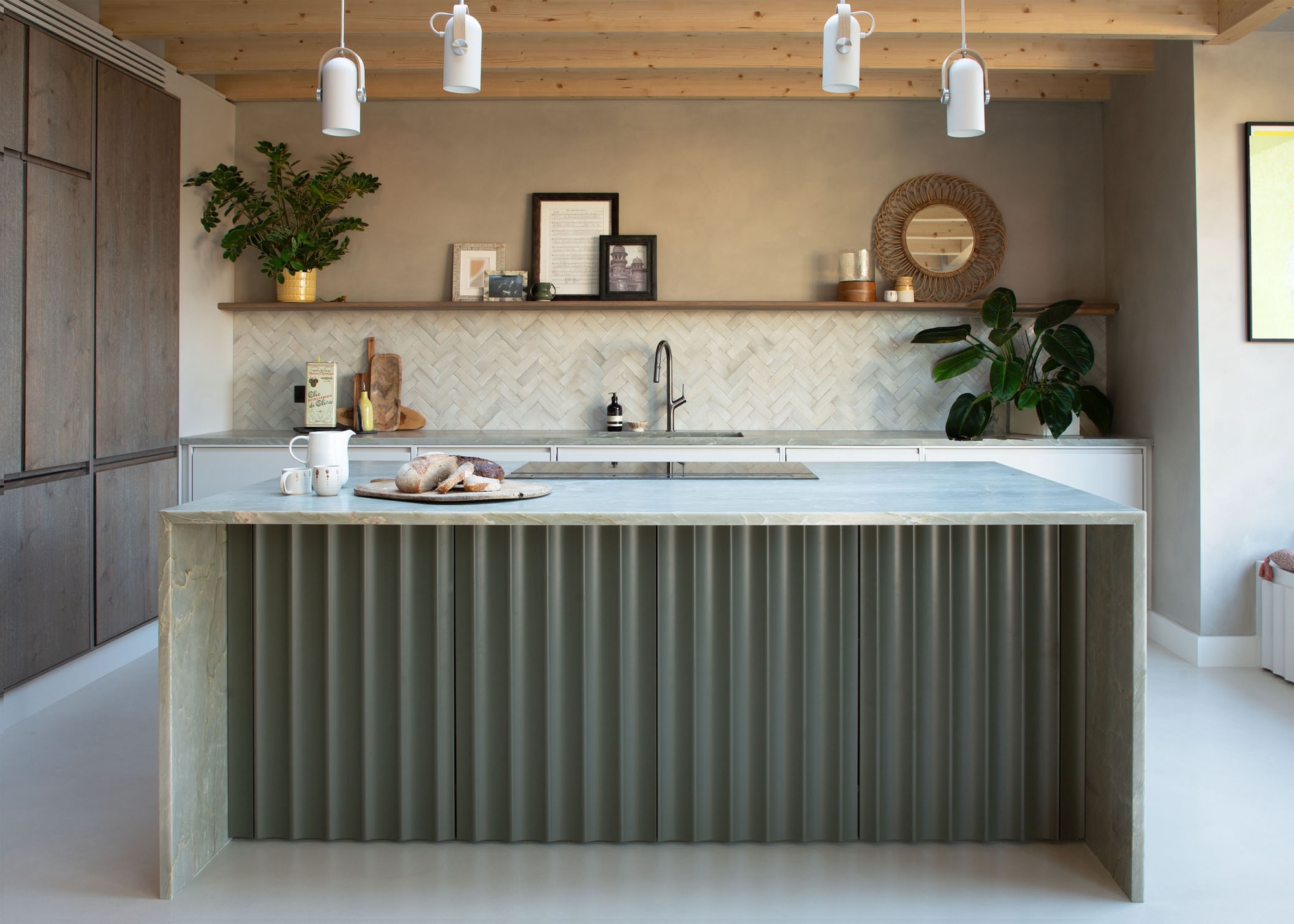 A modern kitchen with a large marble and corrugated metal island, a hob on top and pendant lights above. There are exposed beams on the ceiling and a tiled backsplash.