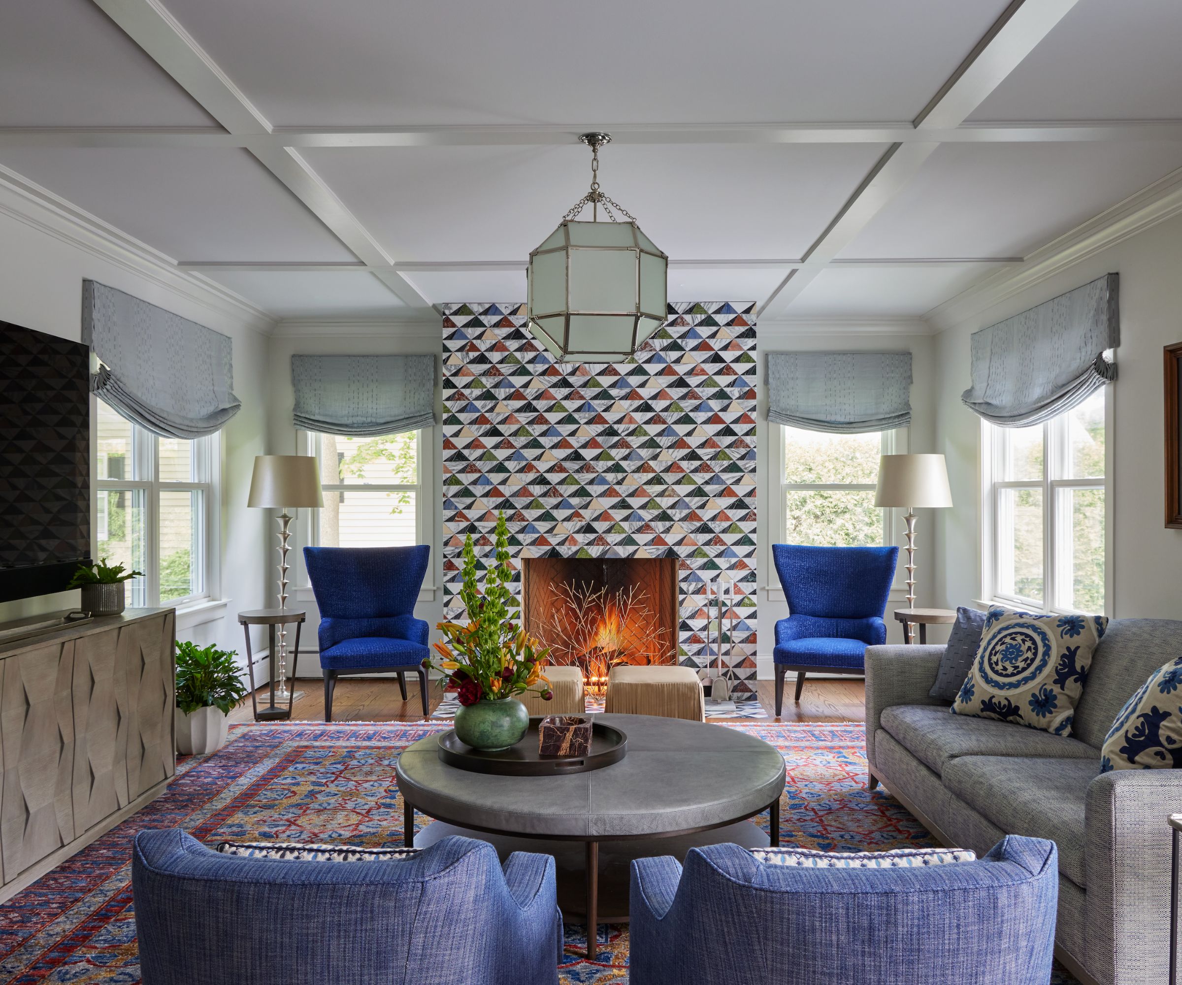 A traditional living room with a cool color scheme, soft gray walls, four blue armchairs, a gray sofa, relaxed blue Roman blinds, a colorful geometric patterned fireplace, and a muted red and blue rug.