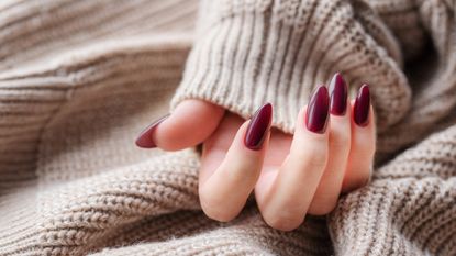 A hand with long, oval shaped dark red nails on a cream, knitted background