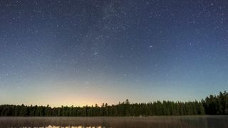 A forest is reflected in a still lake at night with a star filled sky above, part of milky way and andromeda galaxy visible.