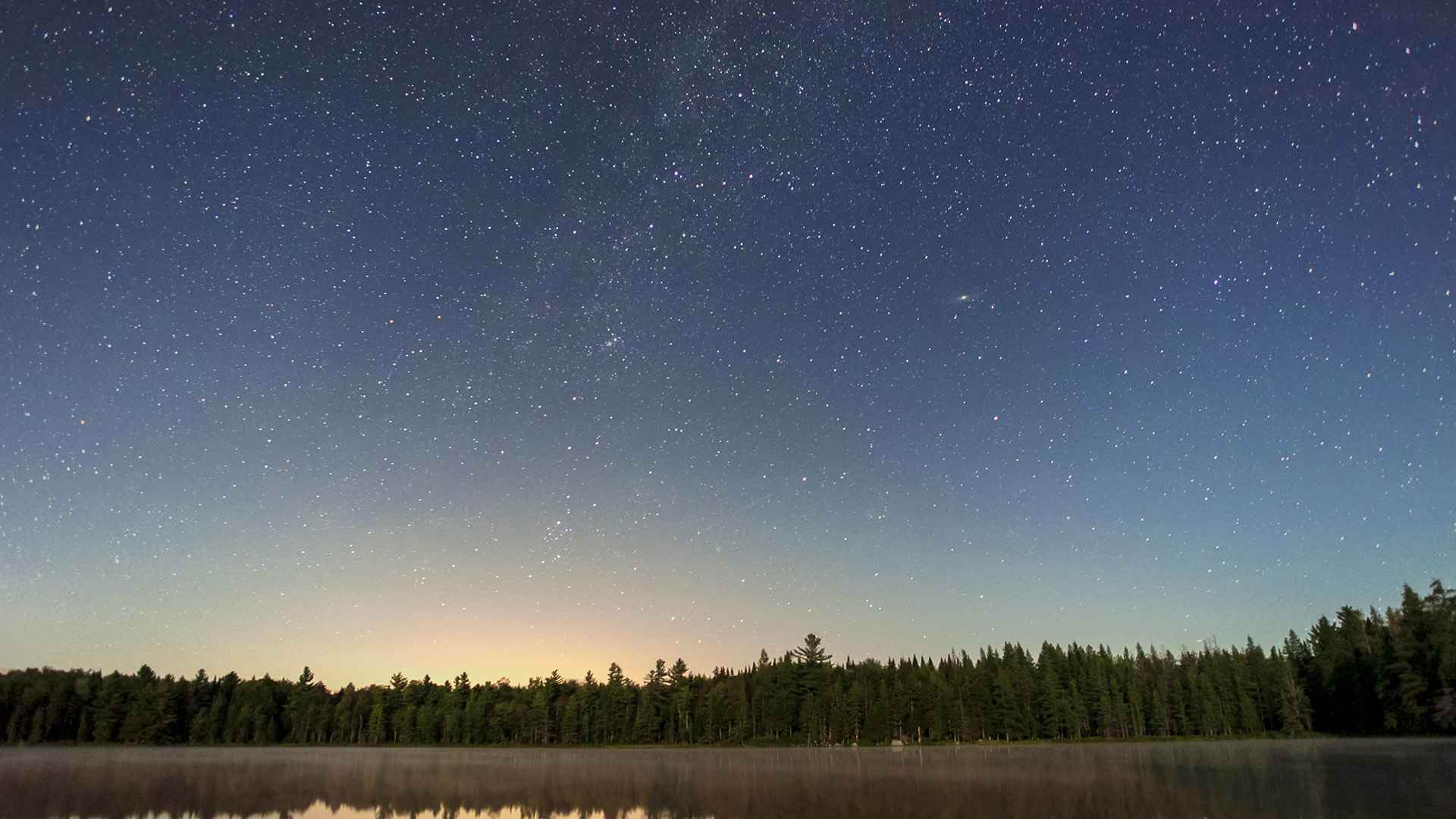 Las odbija się nocą w nieruchomym jeziorze, nad nim rozgwieżdżone niebo, widoczna część Drogi Mlecznej i Galaktyka Andromedy.