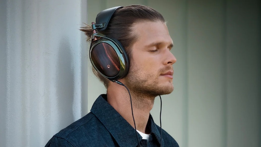 Una foto de perfil de un hombre con los ojos cerrados, con auriculares Meze Strada con cable, apoyado contra un pilar blanco.