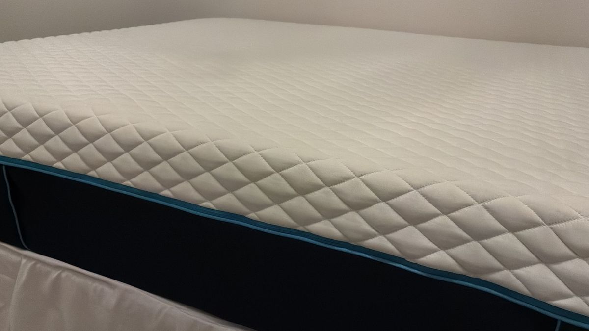 The image shows a white mattress with dark blue sides placed on a mattress base with a grey headboard, set inside a white bedroom