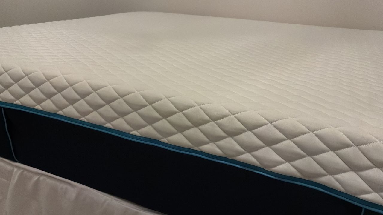 The image shows a white mattress with dark blue sides placed on a mattress base with a grey headboard, set inside a white bedroom