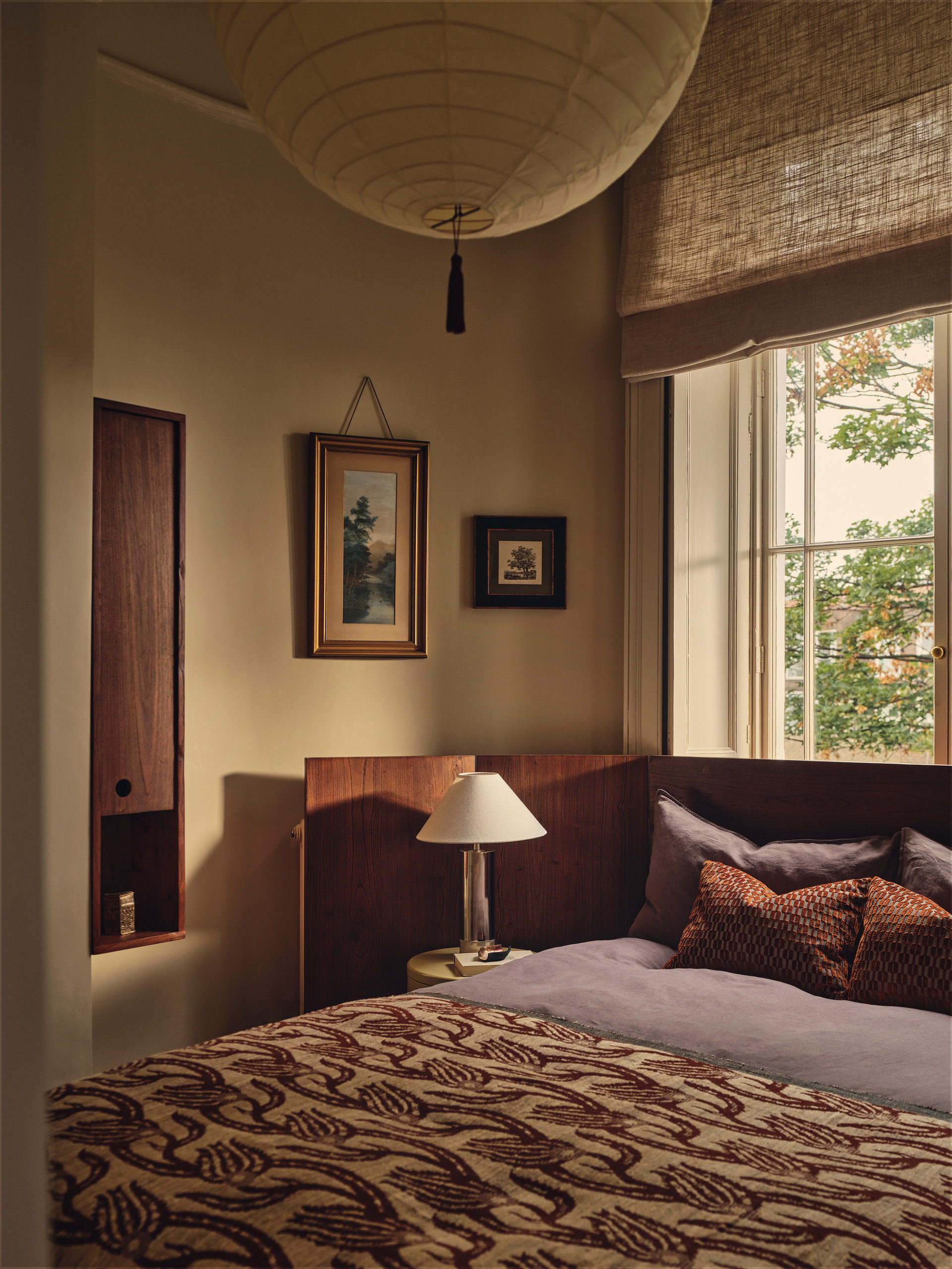Image of an off-white bedroom with a large bed that has an extended wooden headboard. The bedding is purple and red, and there is a lamp by the bed.