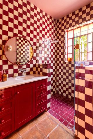 Image of a bathroom covered in tile. The tile is a dark magenta/maroon and white checkered design. The deep magenta color is also in the tile of the shower floor and the cabinetry. There is a round mirror hanging above the sink and the cabinet knobs are round balls.