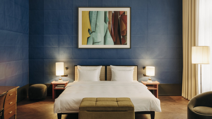 A hotel bedroom with paper-like blue walls with a folded effect, warm bed table lamps and a standing one, green fabric furnishings, wooden pieces, and wooden floors.