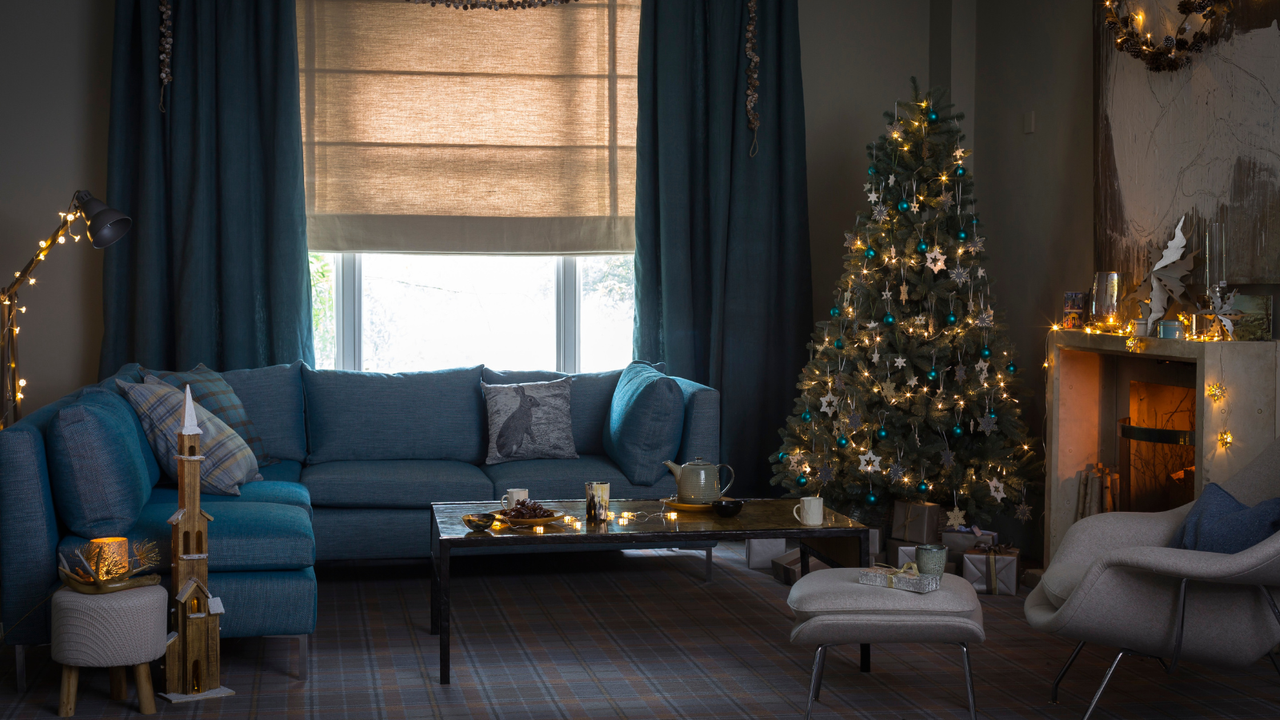 A cozy, festive blue living room with a large blue sofa, decorated Christmas tree and white mantelpiece with a lit fire. In the background is a large window with neutral blinds and blue curtains, and in the foreground is a matching white chair with footrest.