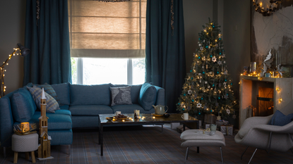 A cozy, festive blue living room with a large blue sofa, decorated Christmas tree and white mantelpiece with a lit fire. In the background is a large window with neutral blinds and blue curtains, and in the foreground is a matching white chair with footrest.