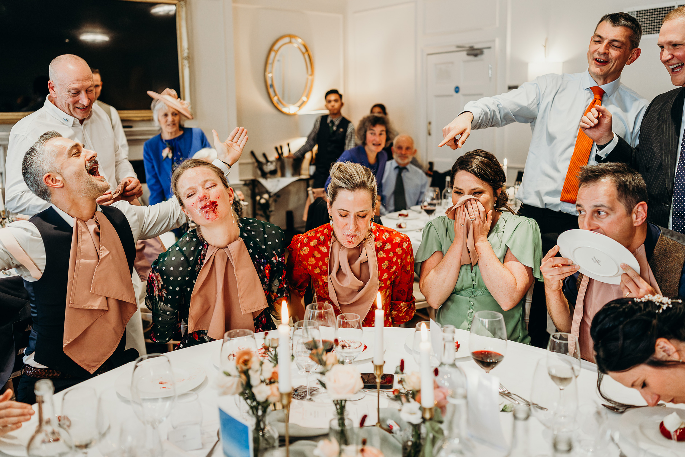 A lively scene at a dining table shows people laughing and playing a game with blindfolds and bibs. Some have food on their faces