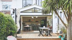 Lea-Wilson house: Rear of house with white extension, wood decking with steps down to grass, outdoor table and chairs