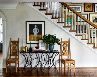 A foyer with neutral mottled wallpaper, a gallery wall or art up the stairs and a long entry table with two chairs