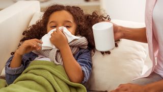 A young girl blows her nose with a cold while a woman holding a mug of tea looks after her