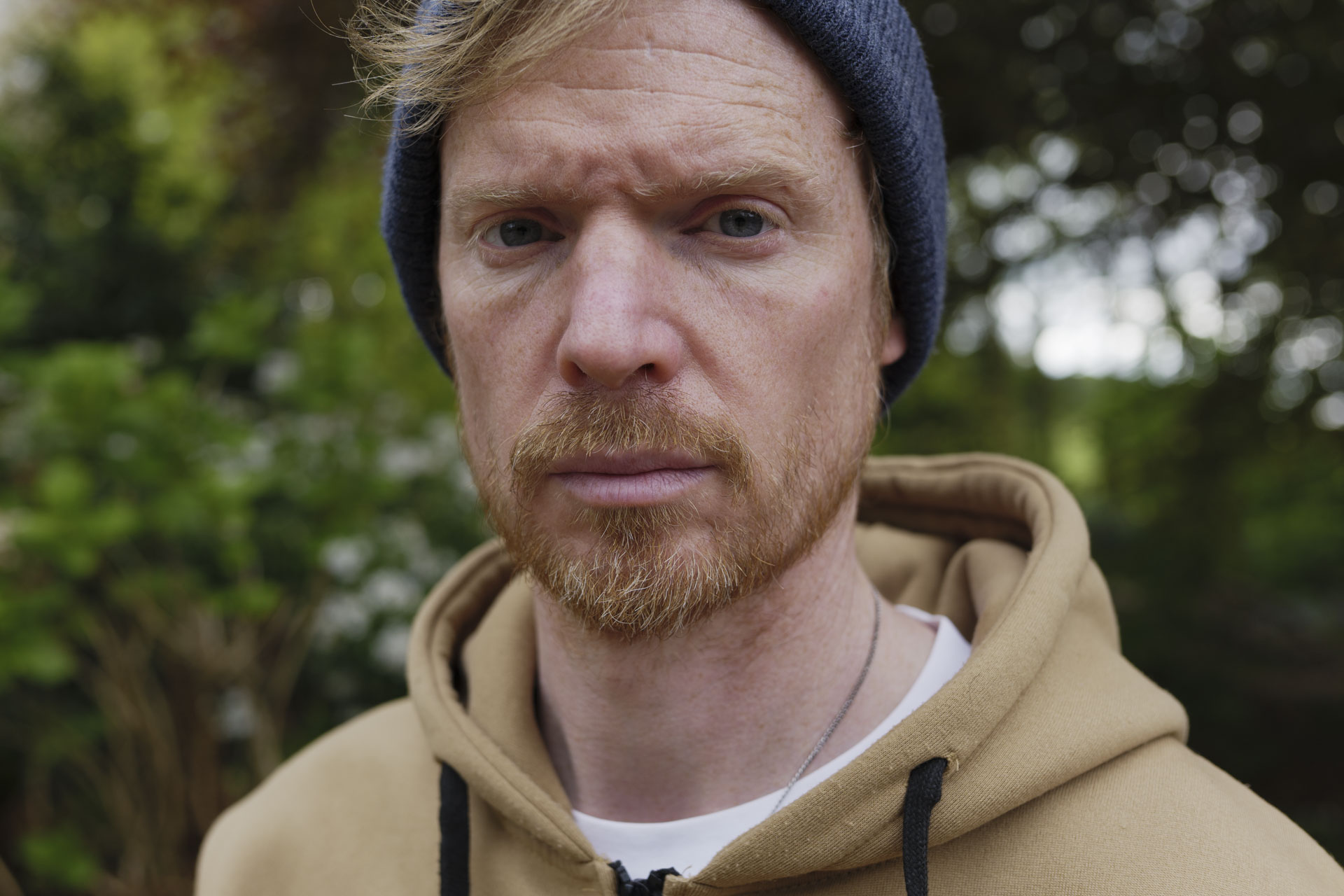 Portrait of a man in a beanie hat and hoodie, the background is lush green bushes and trees with dappled light