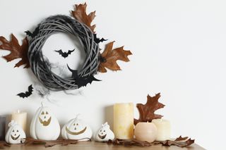 A Halloween wreath made out of wicker hanging on a white wall with bat decorations, candles, and ceramic pumpkins.