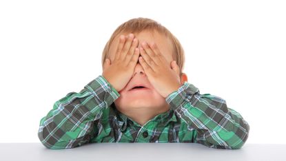Studio portrait of a toddler in a plaid shirt covering his eyes with his hands